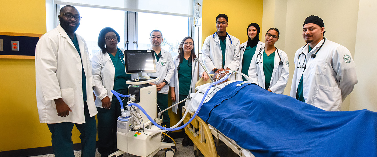 Students in a respiratory therapy lab stand around a patient simulator on a hospital bed, with a ventilator and monitoring equipment in a clinical classroom setting.