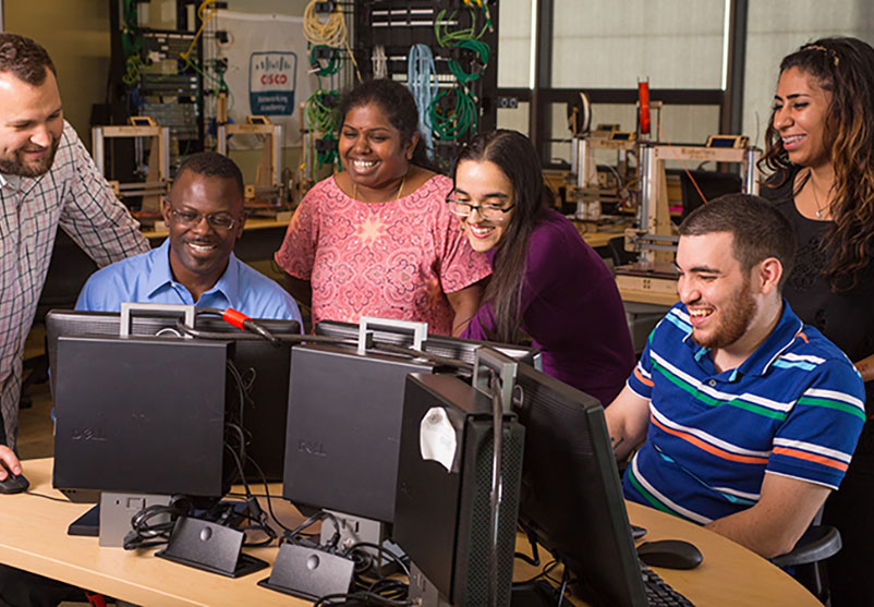 A group of students sitting and standing around two computers smiling.