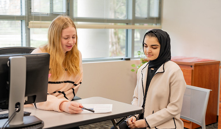 Two women sitting at a desk in an office setting. One is explaining a document while the other listens attentively.