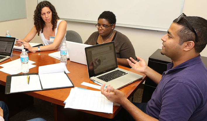 Group of three women and one man collaborating at a table with laptops, notebooks, and water bottles in a meeting room.