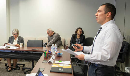 Man in a white shirt presenting in a conference room, holding papers. Two colleagues sit and listen in the background.