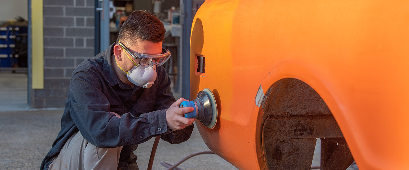 Student sanding an orange car in a repair shop.