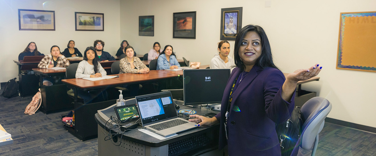 Instructor gestures toward students during a classroom lecture while seated at a computer workstation, with students listening and taking notes in the background.