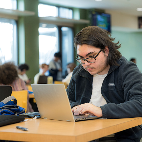 Student working on a laptop in a busy campus common area, focused on their screen with a backpack and notebook nearby.