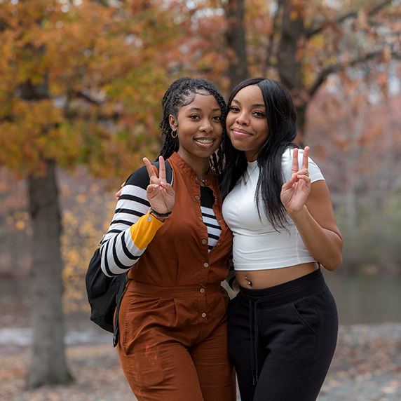 Two students pose outdoors in front of autumn trees, smiling and holding up peace signs.