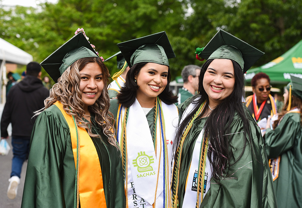Three graduates in green caps and gowns smiling together outdoors, wearing honor cords and stoles during a commencement ceremony.